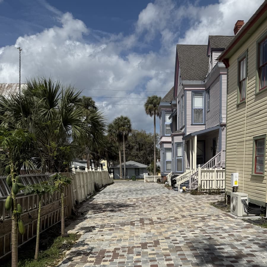 A paved pathway flanked by colorful houses and palm trees under a partly cloudy sky.