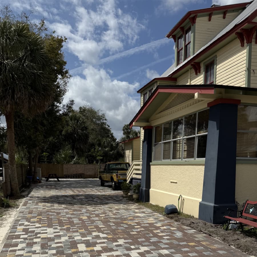 A colorful paved driveway leads up to a large, two-story house under a partly cloudy sky.