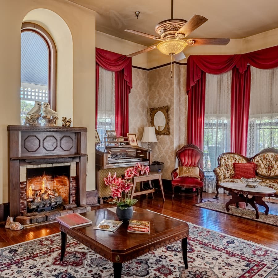 A vintage living room featuring a fireplace, musical instrument, elegant seating, and floral decor.